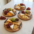 Three round rustic wooden serving trays with black metal handles styled with charcuterie on a white marble surface